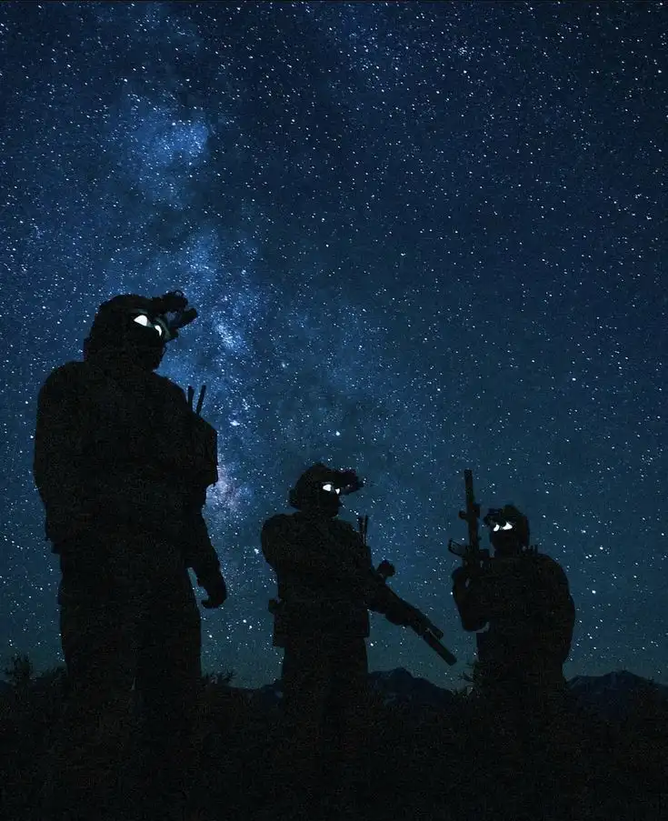 Three silhouetted soldiers in tactical gear stand against a backdrop of a star-filled night sky, their helmet lights…