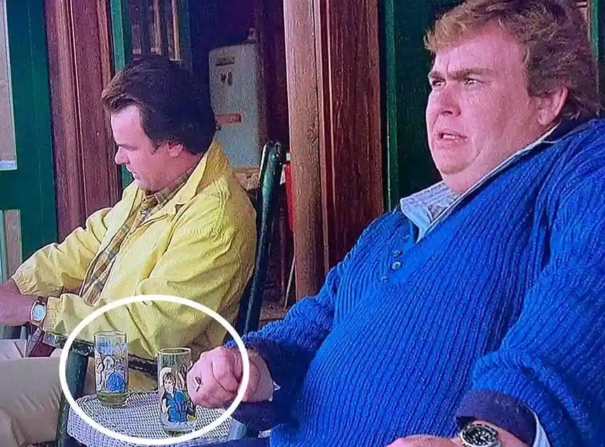 two men sit at a table; the man in the blue sweater has a disgusted expression while the man in the yellow jacket looks…
