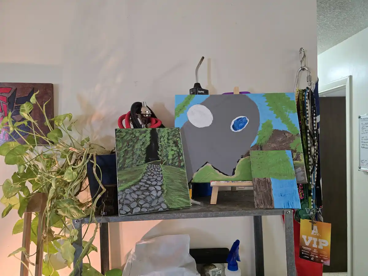 A cluttered metal shelf holds several unfinished paintings, including one of a large gray skull with a white and blue eye…
