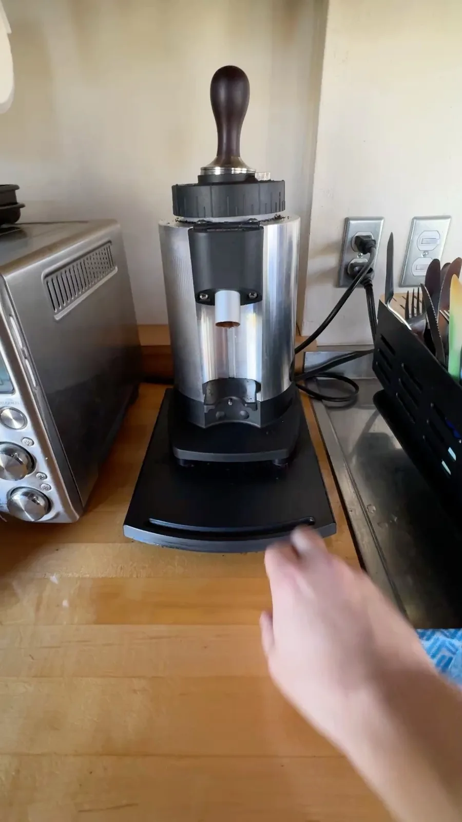 A hand reaches toward a stainless steel coffee grinder with a wooden handle, sitting on a wooden countertop next to a…
