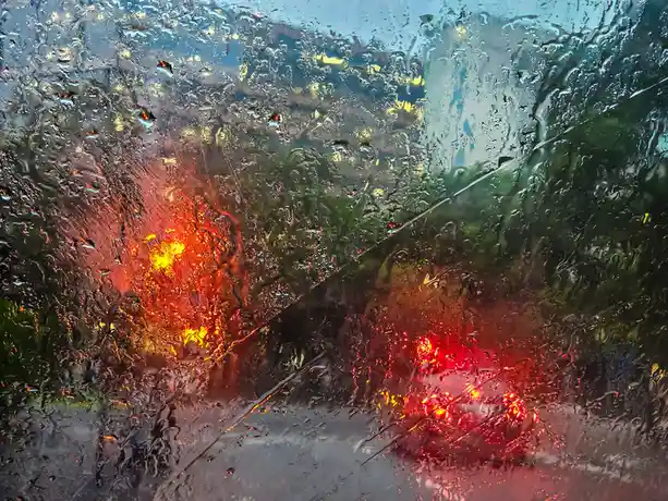 A view from inside a car looking out through a rain-splattered windshield, with blurred red and yellow lights from…