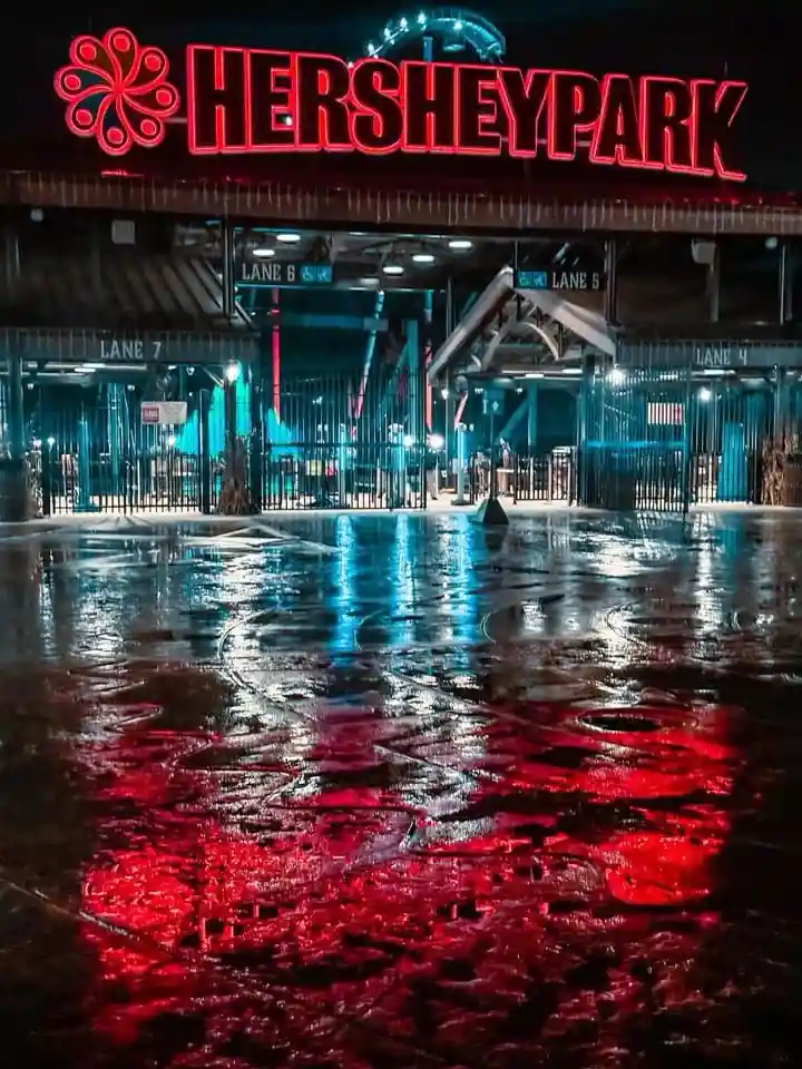 A nighttime photo of the Hersheypark entrance with a large red neon sign above gated lanes, all reflected in wet pavement…
