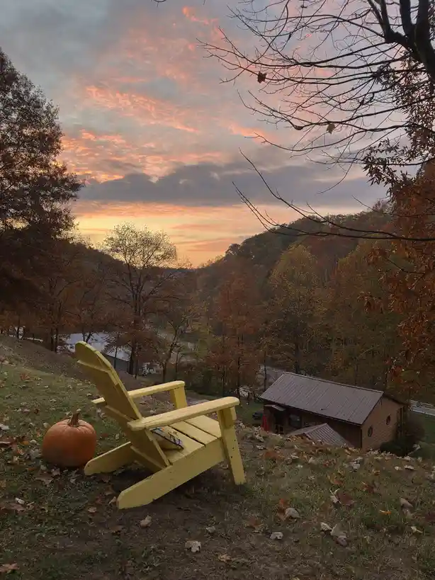 A bright yellow Adirondack chair sits on a grassy hillside next to a pumpkin, overlooking a valley with autumn-colored trees…