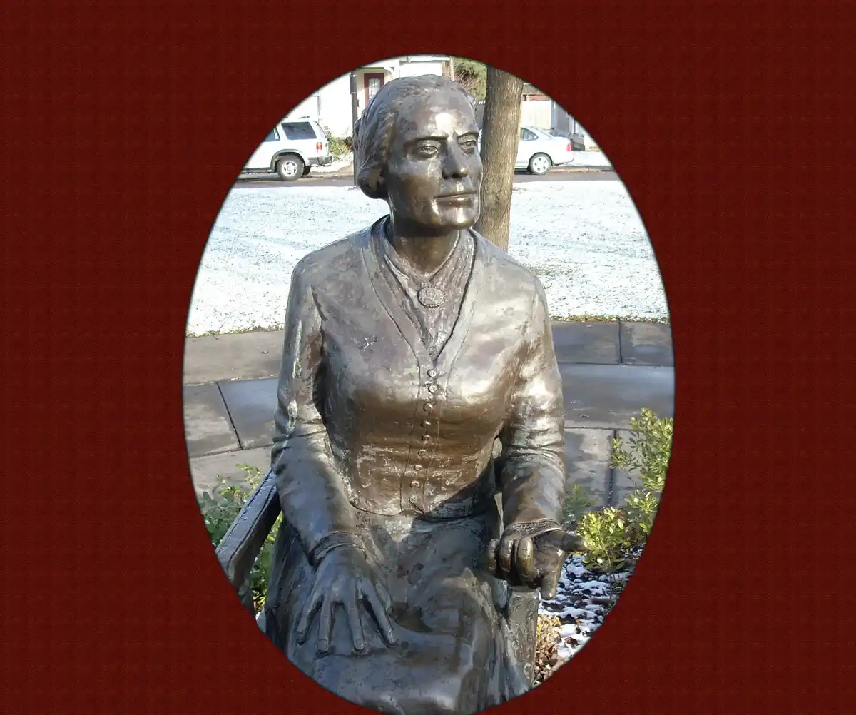 A bronze statue of a seated woman in period clothing is framed within a circular cutout against a textured red background…