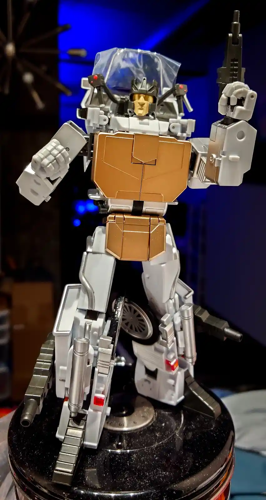 A detailed transformer toy, mostly white and gold, stands on a black circular base with its right arm raised holding a gun…