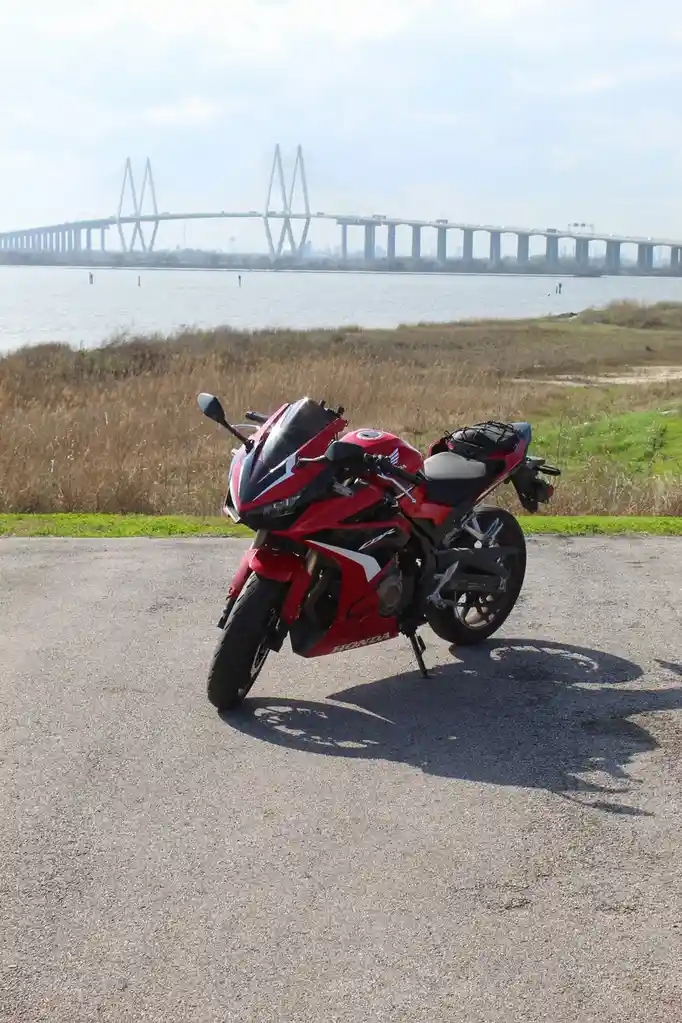 A red Honda CBR motorcycle is parked on an asphalt path near a body of water, with a large cable-stayed bridge visible in…