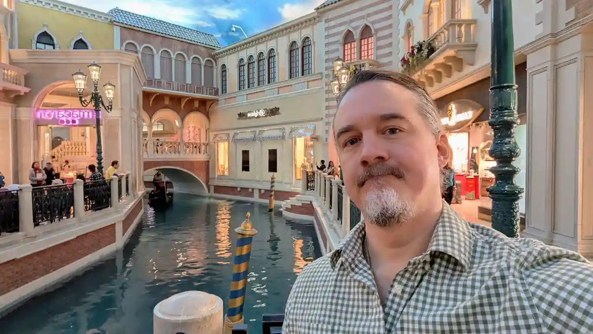 A man with a graying beard and mustache wearing a green and white checkered shirt takes a selfie in front of a replica…