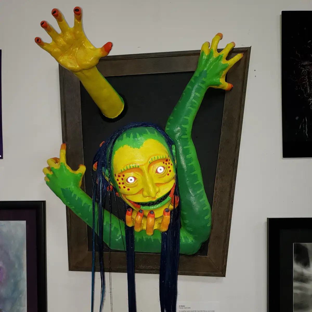 A three-dimensional sculpture of a grotesque, smiling creature with green and yellow skin, blue hair, and glowing white…