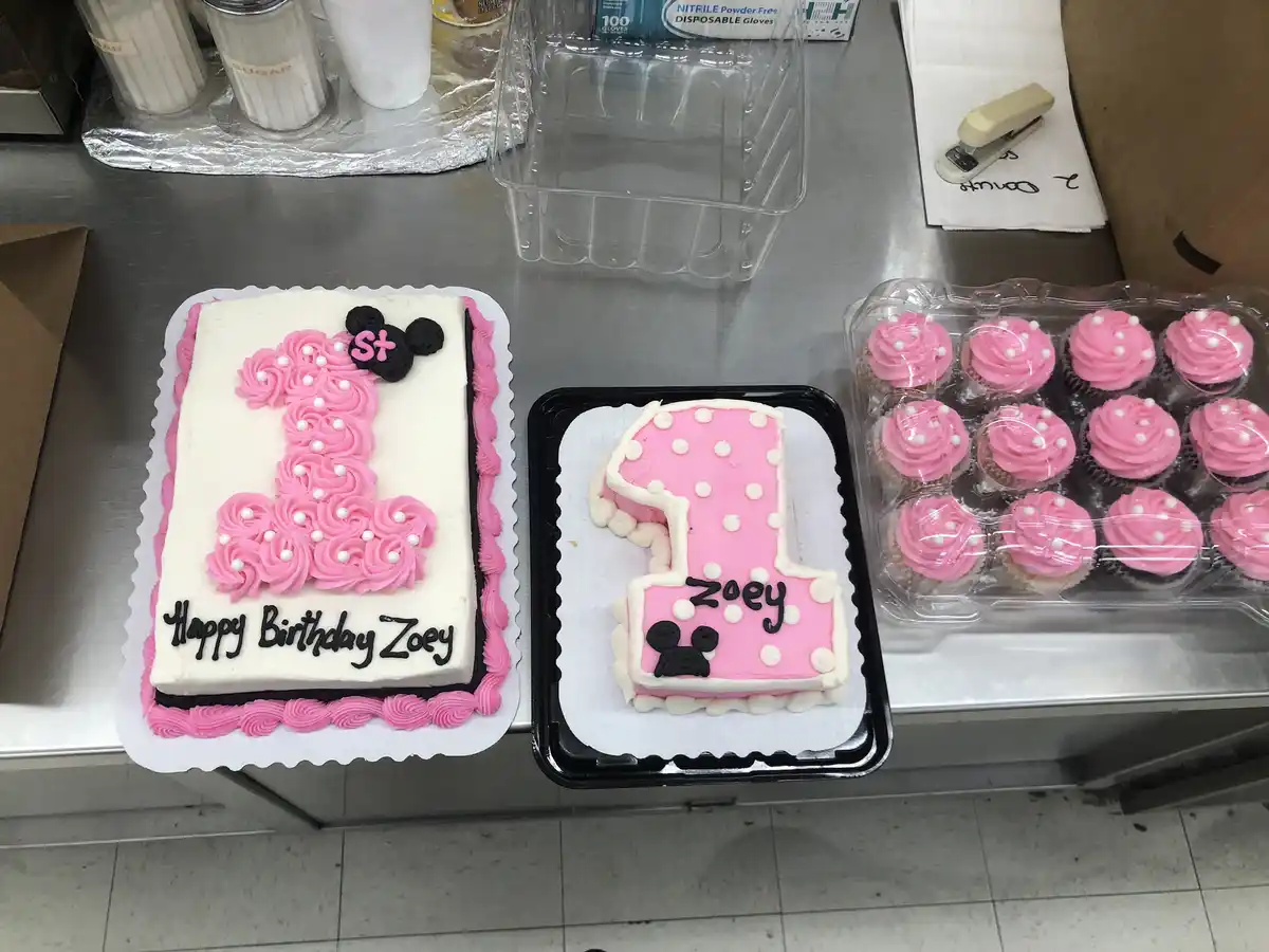 Two identical first birthday cakes for 'Zoey' are displayed side-by-side on a stainless steel counter, both shaped like the…