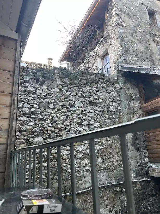A low-angle shot from a balcony shows a weathered stone wall between old buildings, with a metal railing in the foreground…