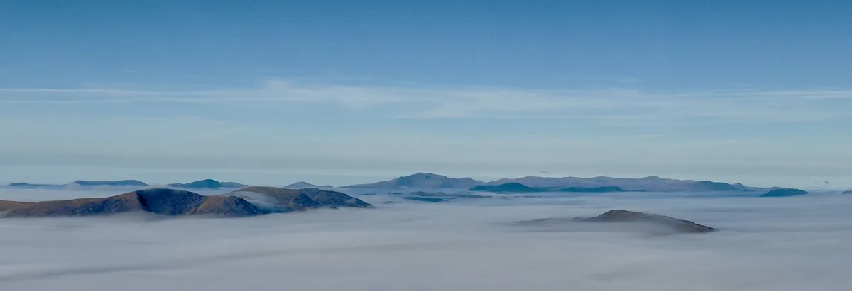 A serene landscape photo shows rolling mountains partially emerging from a thick, soft blanket of clouds that fills the…