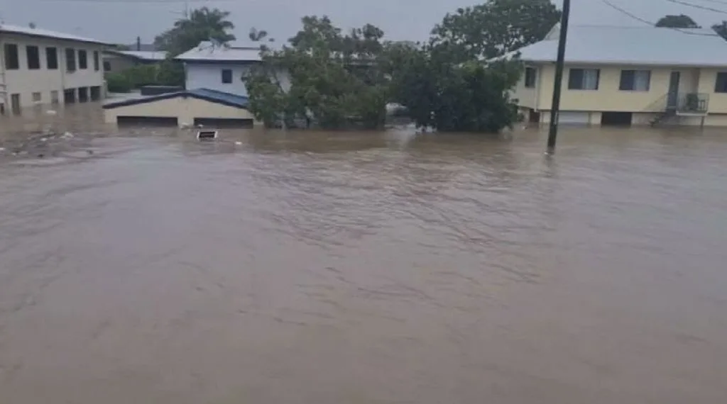 A flooded residential street with murky brown water covering the road and reaching halfway up the walls of single-story…
