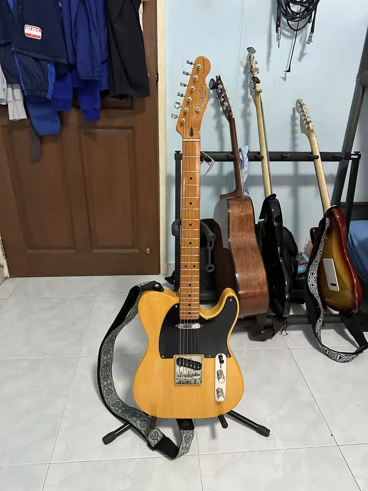 A light-wood finish Squier Telecaster electric guitar stands upright on a stand in a room, with a patterned strap draped…