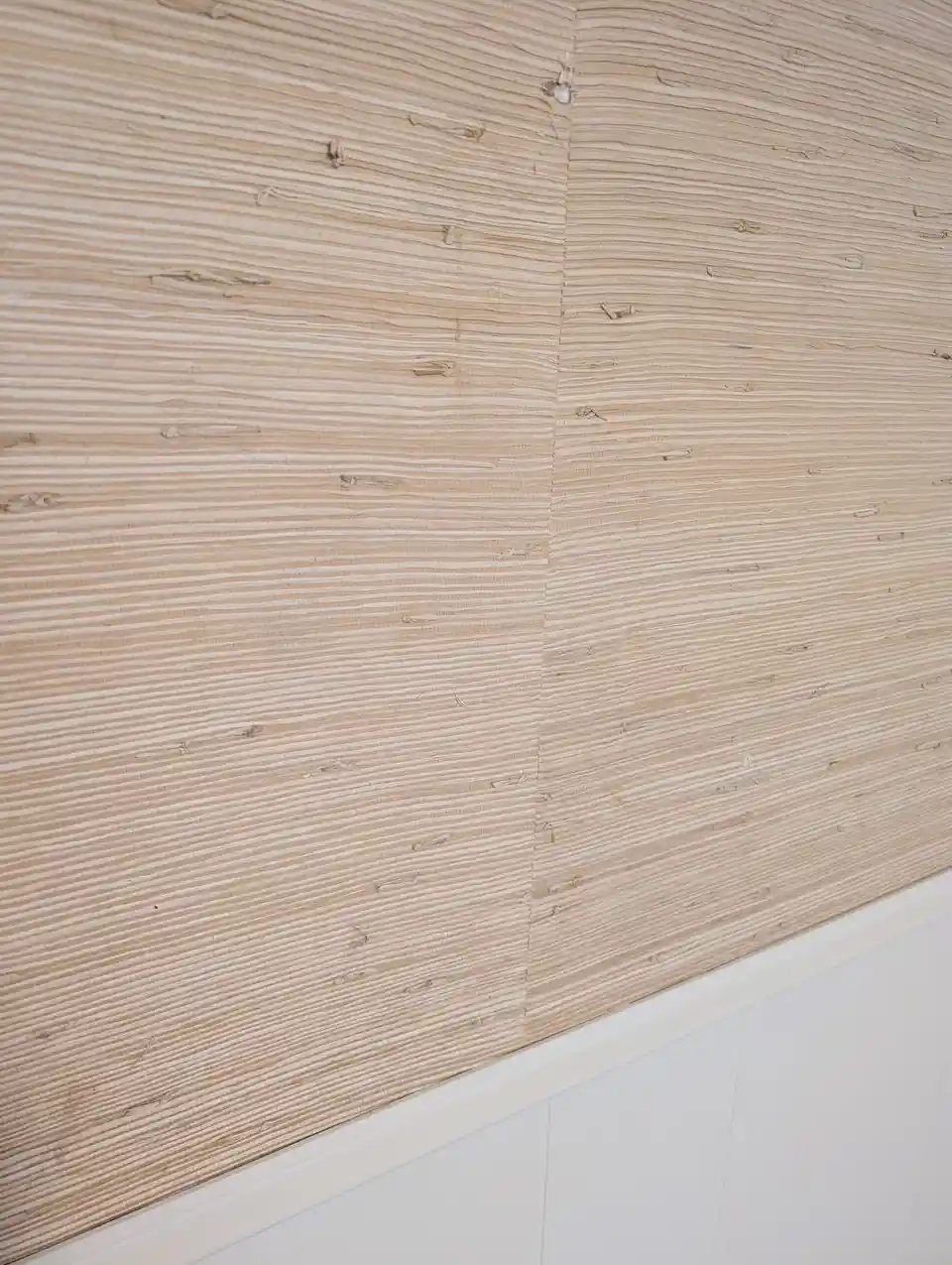 A close-up photo shows a wall covered in light beige textured wallpaper with horizontal ridges and small imperfections…