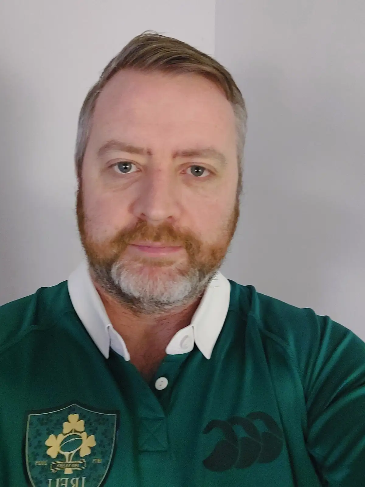A man with a beard and grayish hair wears a green Irish rugby jersey with a shamrock logo and a black three-leaf clover…