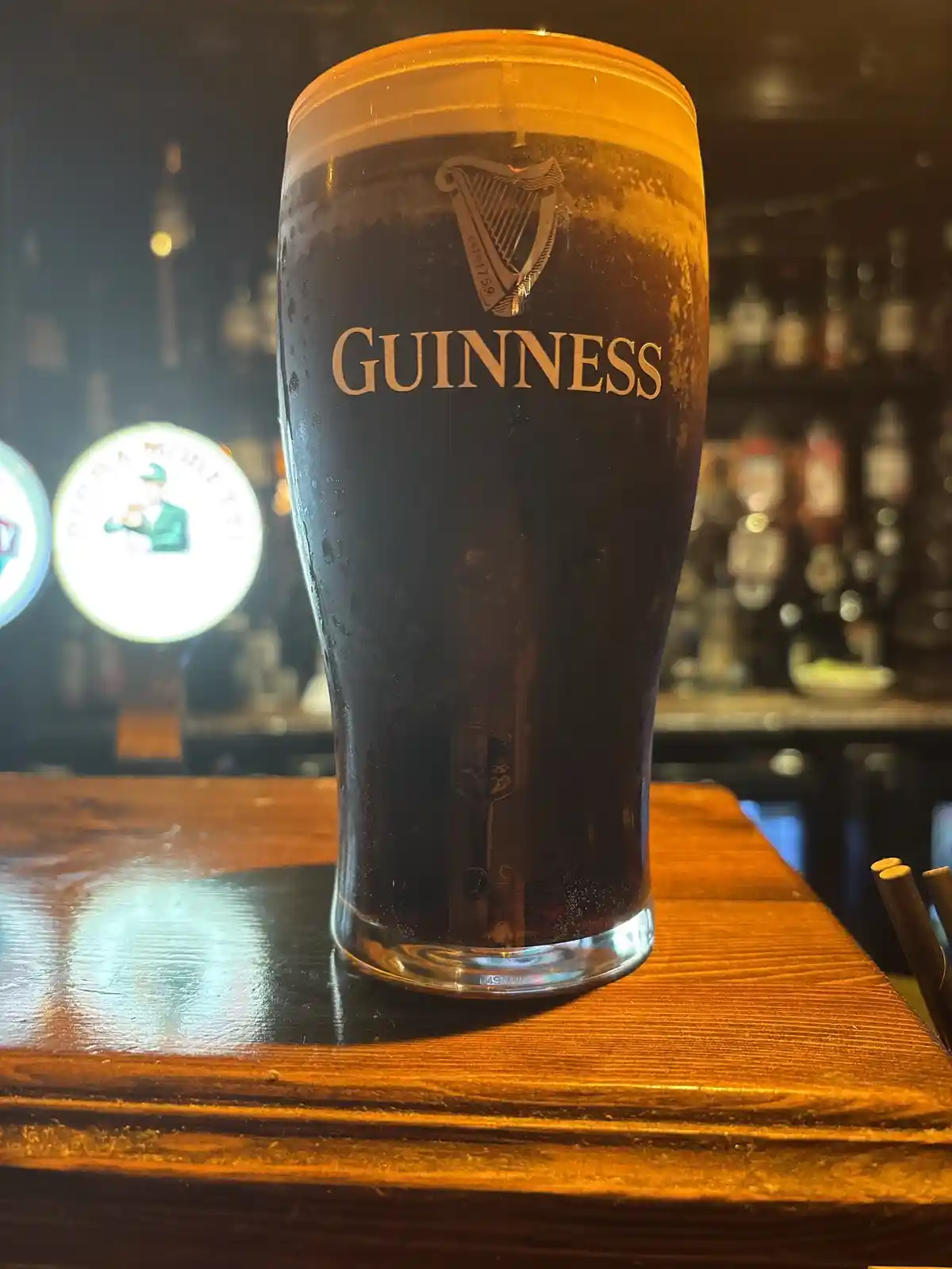 A tall, chilled pint of Guinness beer sits on a wooden bar top, its dark liquid topped with a creamy tan head. The iconic…