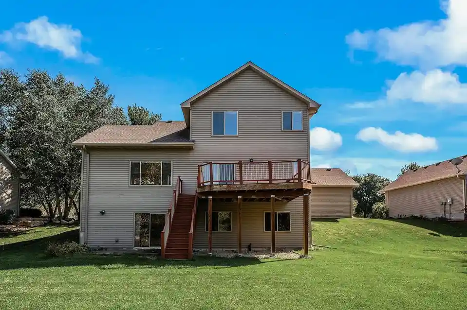 A two-story beige house with a wooden deck and stairs, set against a bright blue sky with fluffy clouds and green grass. The…