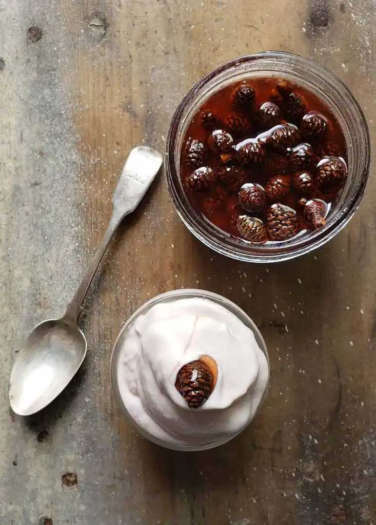 An overhead shot shows a glass jar of white yogurt topped with a single pinecone, next to a glass jar filled with pinecones…
