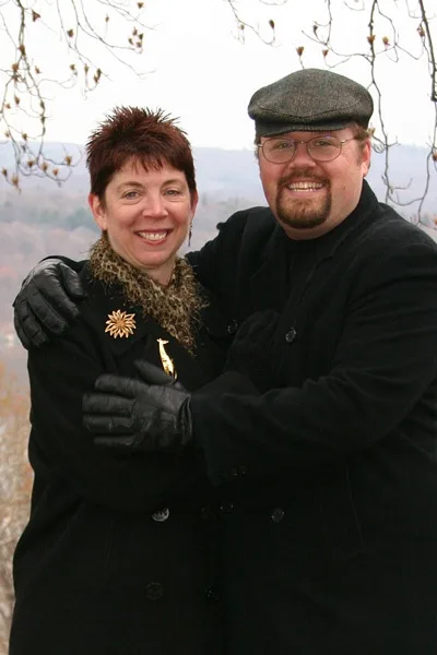 A smiling man and woman pose outdoors in winter coats; the man has his arm around the woman, who wears a black coat with a…