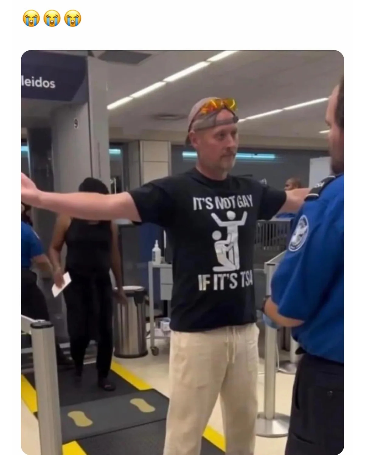 A man at an airport security checkpoint, arms outstretched like he's being scanned, wears a black t-shirt with white text…