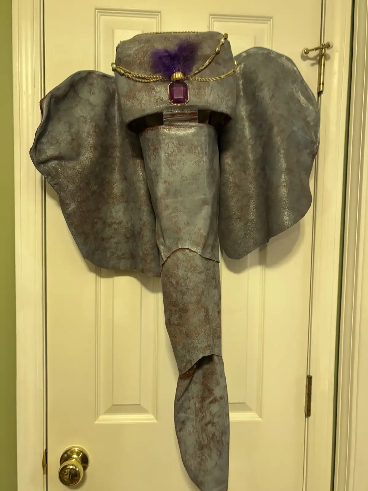 A large, gray, elephant-shaped doorstop with a textured, faux-leather surface and a purple jewel and feather headpiece is…
