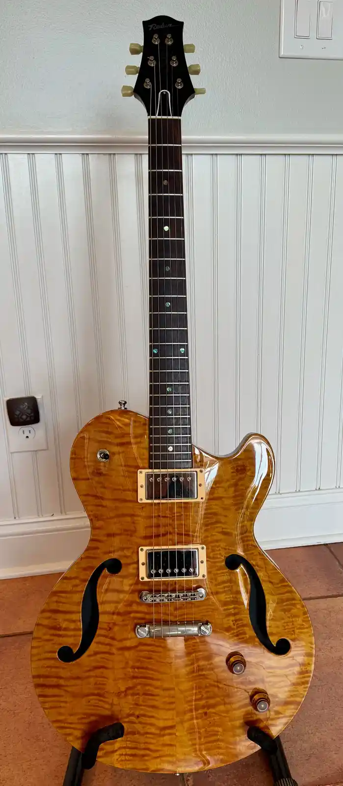 A full-body photo of a glossy, amber-finished electric guitar with a distinctive double-cutaway body and f-holes, standing…