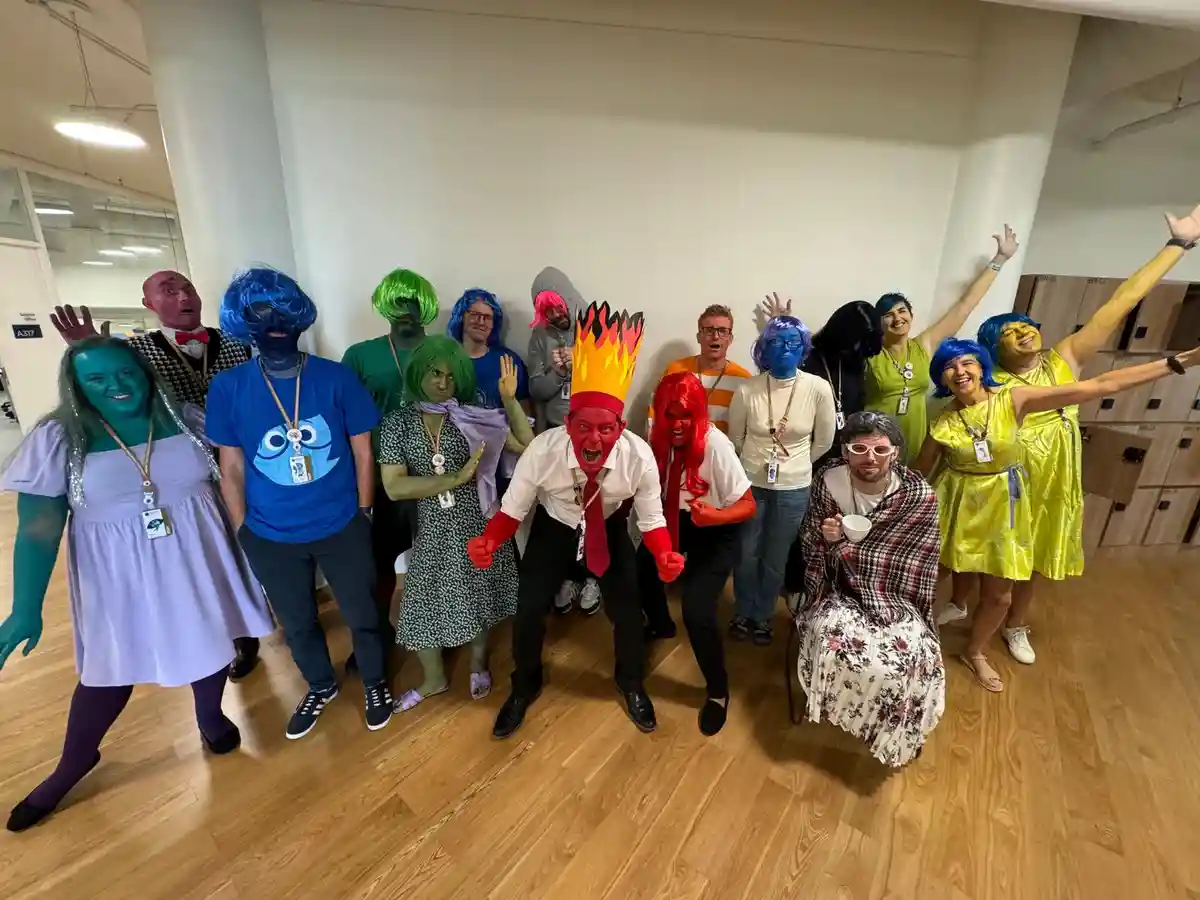 A group of office workers in a brightly lit room are dressed as colorful characters from the animated show 'The Amazing…