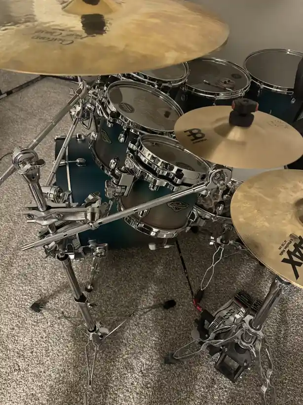 A high-angle photo shows a complete drum set with chrome hardware, teal-colored drums, and brass cymbals resting on a…