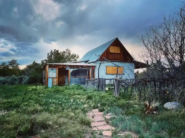A dilapidated, weathered house with boarded-up windows and a rusted roof sits in a grassy field under a dramatic, cloudy…