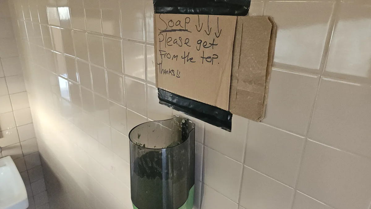 A cardboard sign taped to a tiled bathroom wall above a nearly empty soap dispenser, handwritten in black marker with arrows…