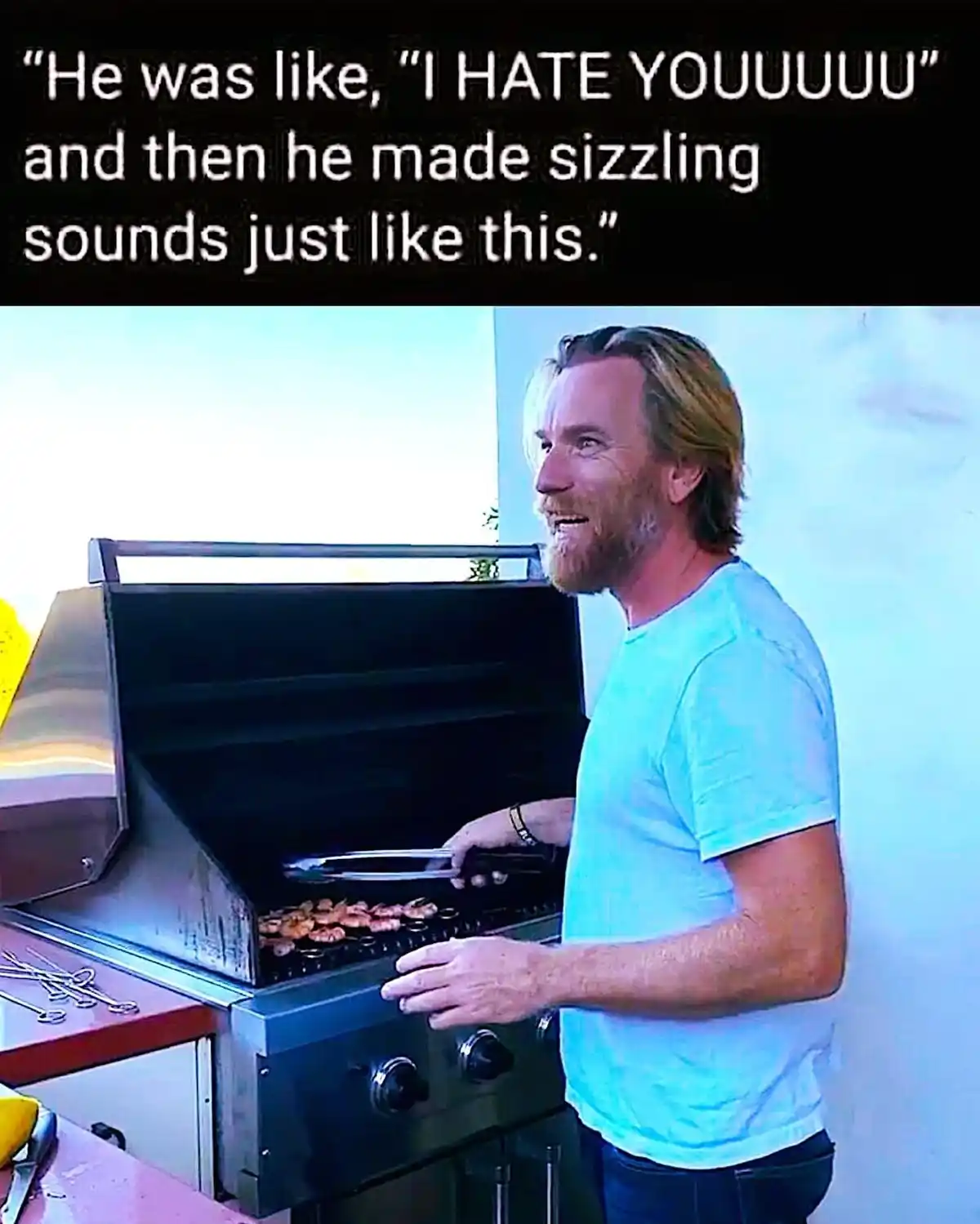 A man with a beard and light blue t-shirt stands beside an open stainless steel grill, holding tongs over shrimp cooking on…