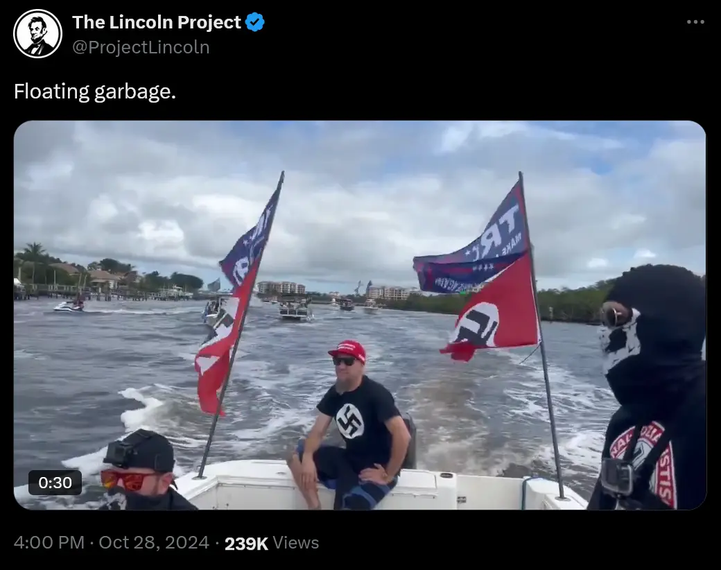 A boat with several people, including one wearing a red cap and a black t-shirt with a swastika, is shown on water with…