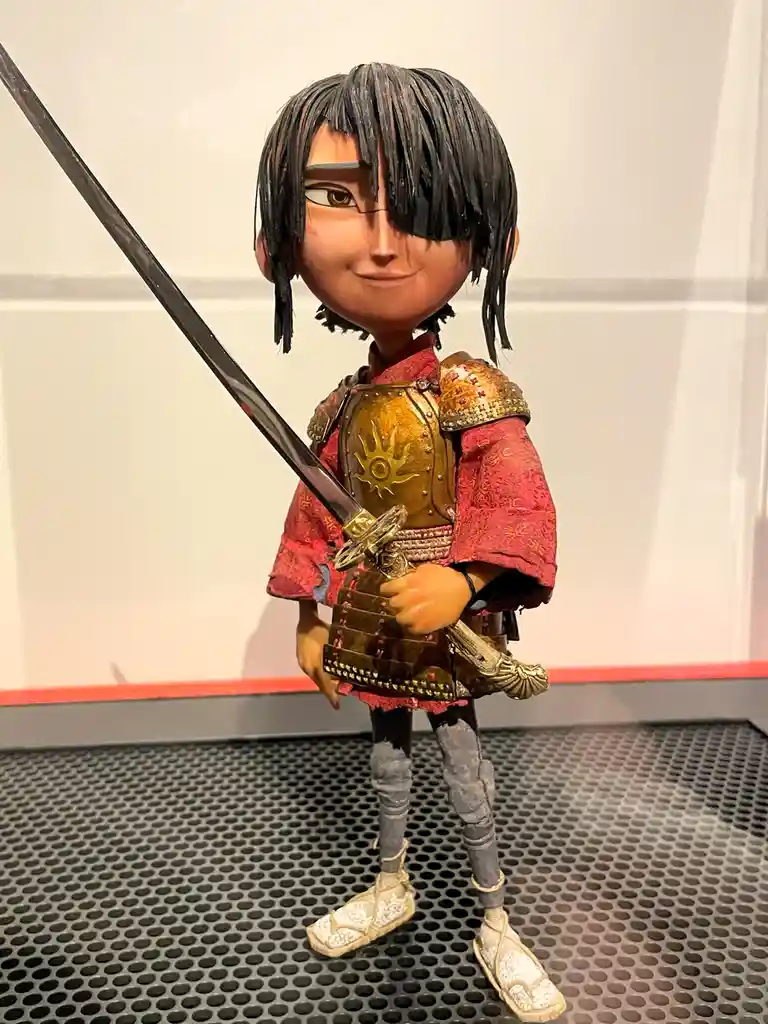 A figurine of the animated character Haku from 'Mulan' is shown standing on a perforated floor, holding a sword. He has a…