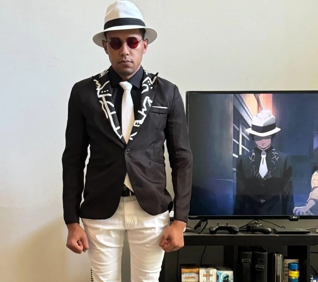 A man wearing a white fedora, red-tinted sunglasses, a black blazer with white abstract embroidery, a white tie, and white…