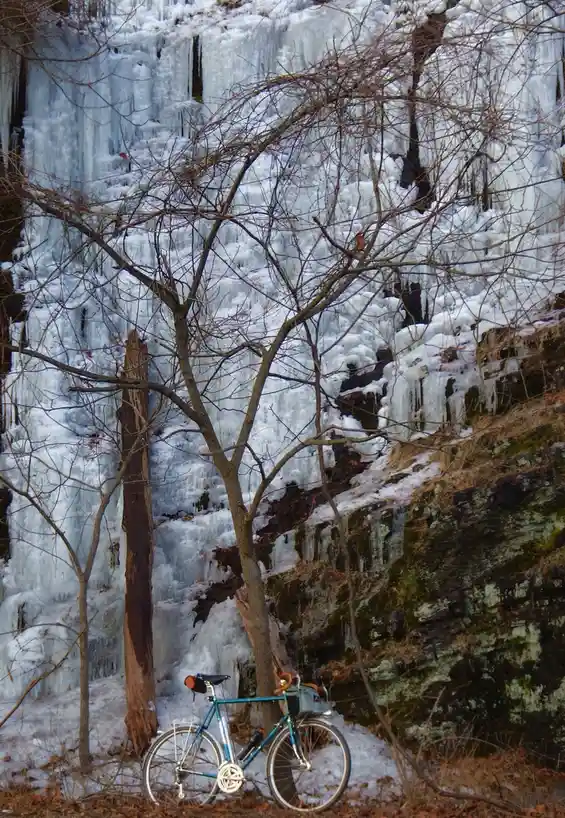 A teal bicycle is parked in front of a large frozen waterfall covered in thick ice and icicles, with bare trees in the…
