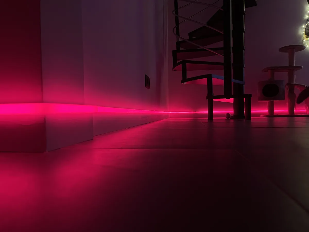 A low-angle shot of a dimly lit room bathed in vibrant pink LED strip lights along the baseboards. Visible are a black…