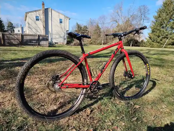 A red mountain bike with black tires and a black saddle is parked on grass in a backyard, with a house and trees in the…
