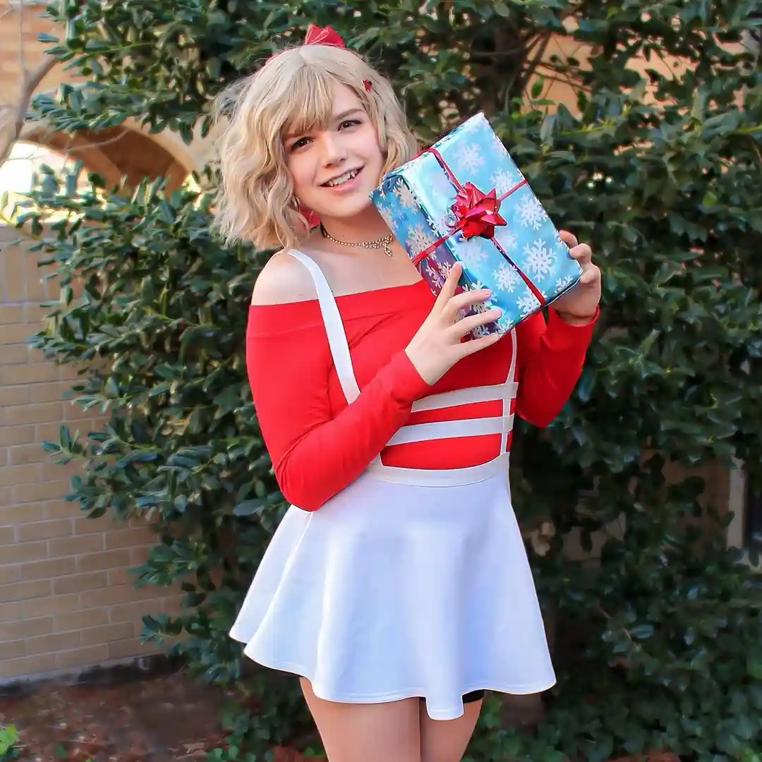A person with blonde hair and a red bow holds a large blue gift wrapped in snowflake paper with a red bow. They wear a red…