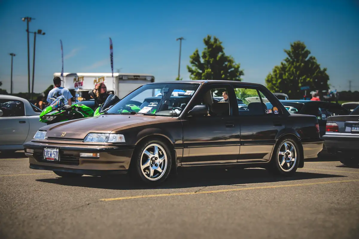 A brown Honda Accord sedan is parked in a sunny outdoor car show lot, surrounded by other cars and people. The car has…