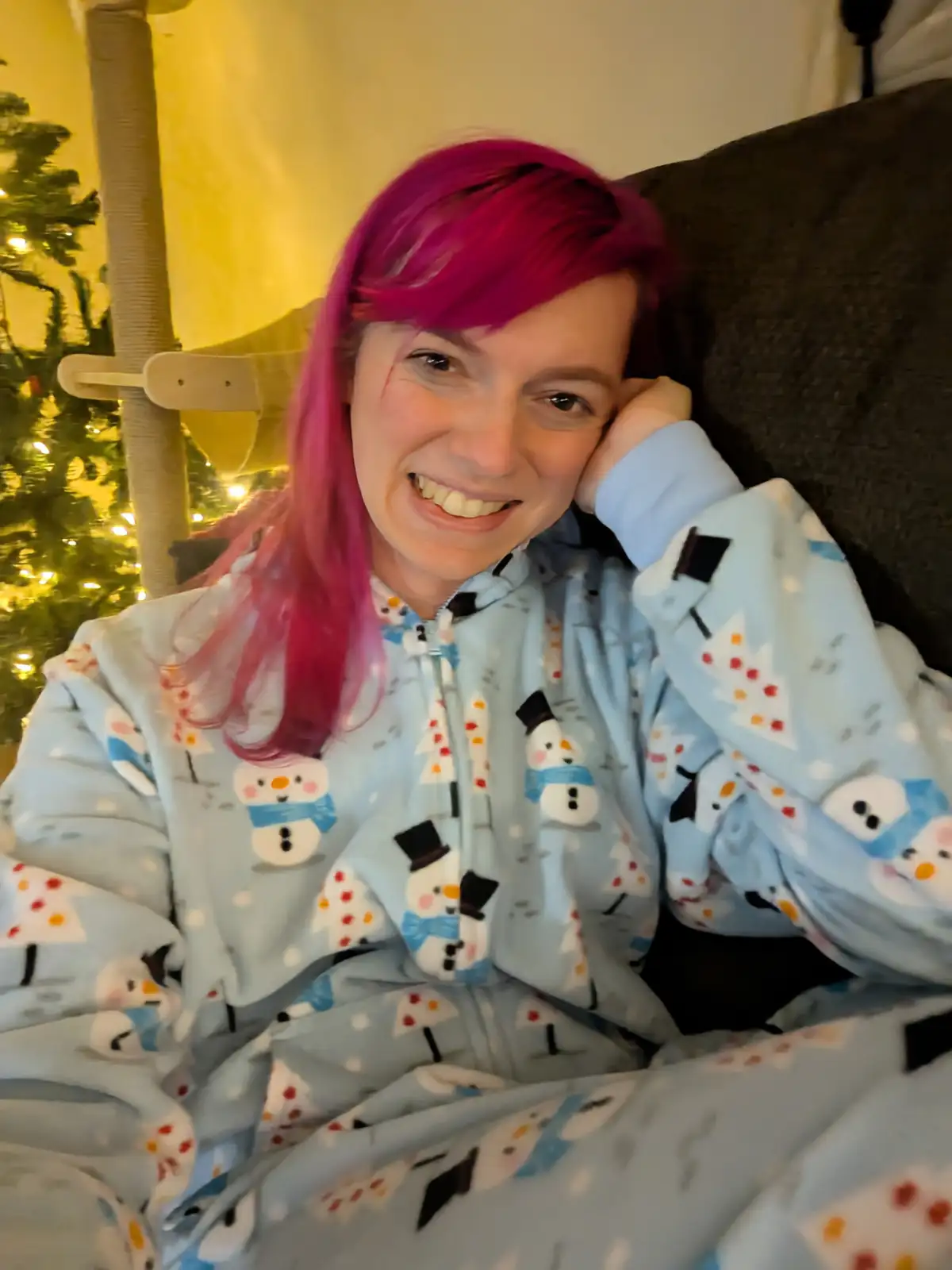 A person with bright pink hair is smiling while lounging on a couch, wearing light blue pajamas covered in snowman and…