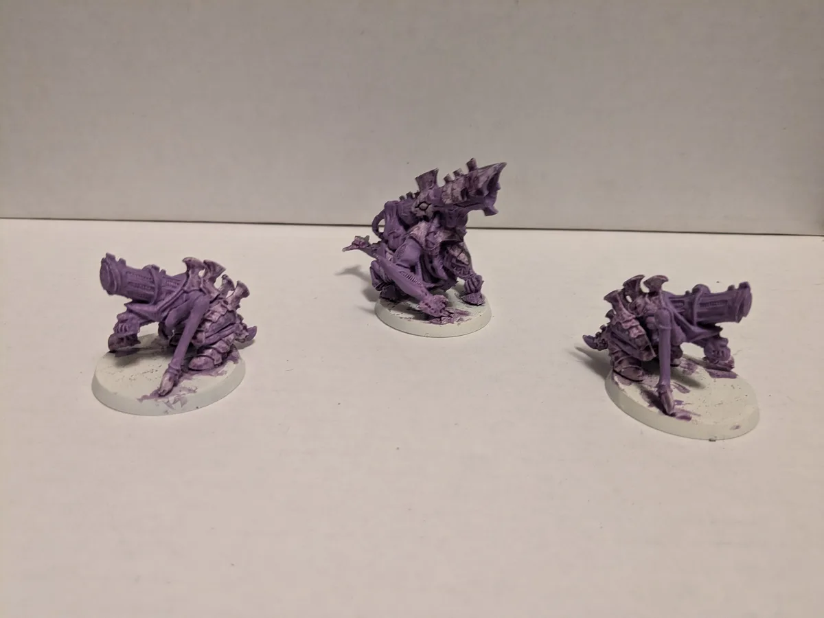 Three unpainted purple fantasy miniatures on white circular bases are arranged on a plain white surface. The central figure…