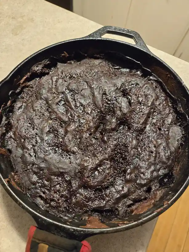 A close-up photo shows a dark, glossy, slightly cracked chocolate brownie baked in a black cast-iron skillet resting on a…
