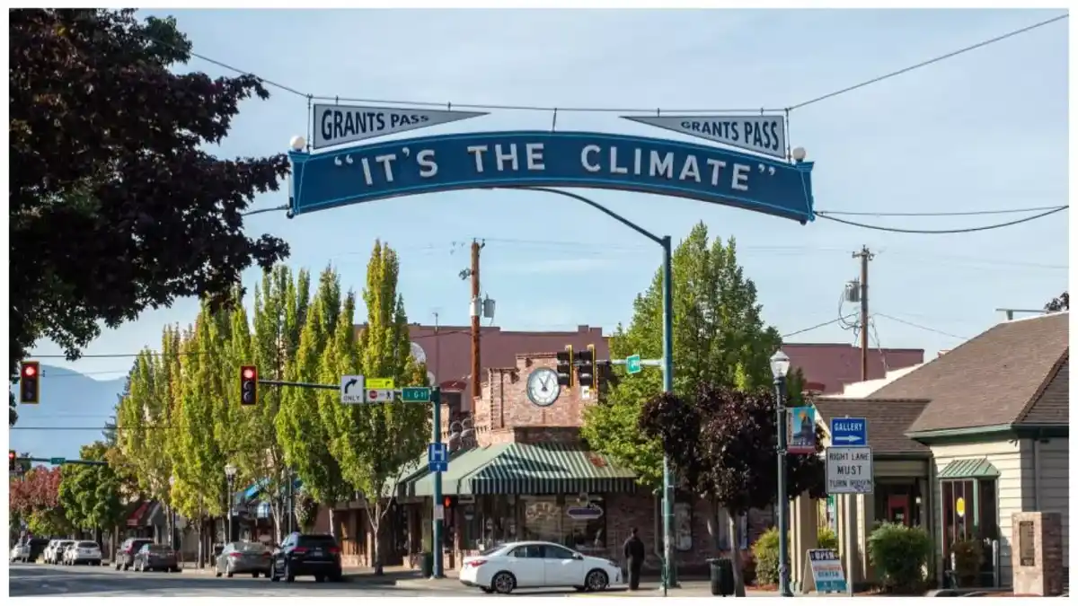 A street view of Grants Pass, Oregon, featuring a large blue archway sign spanning the road with the text 'IT'S THE CLIMATE'…