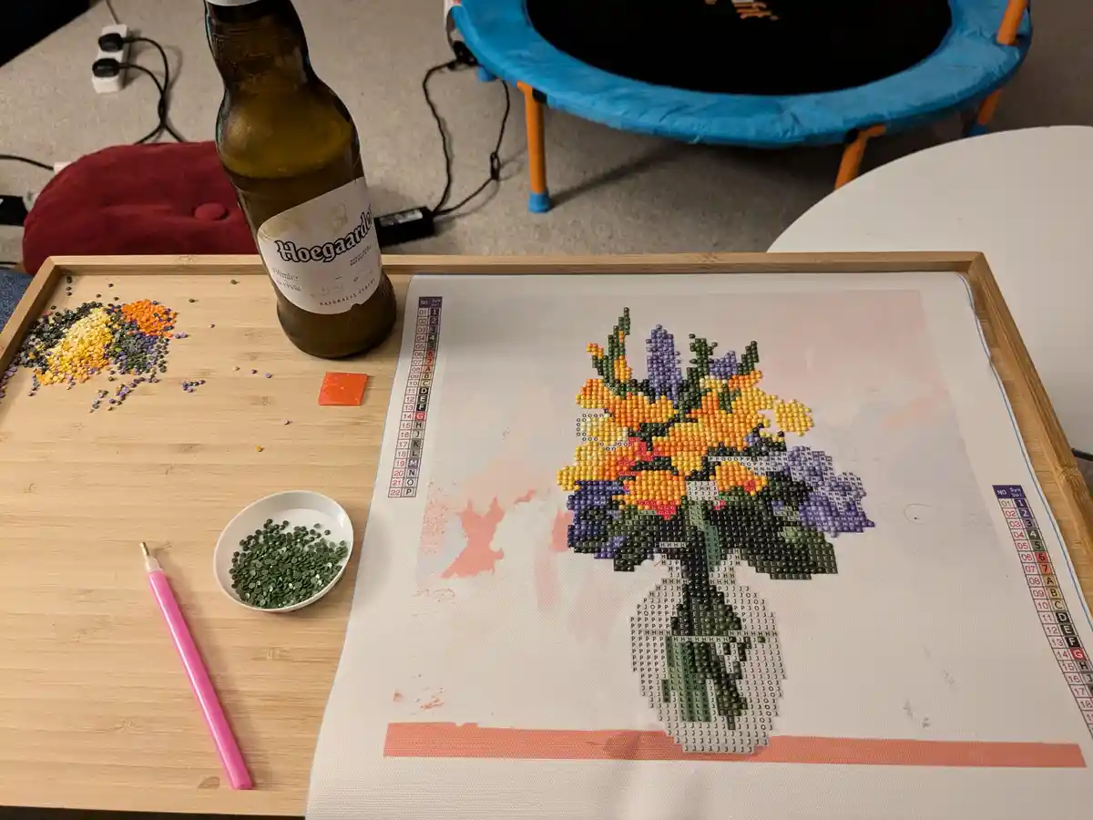 A partially completed bead art project of a flower bouquet on a canvas sits on a wooden tray, with spilled beads, a pink…