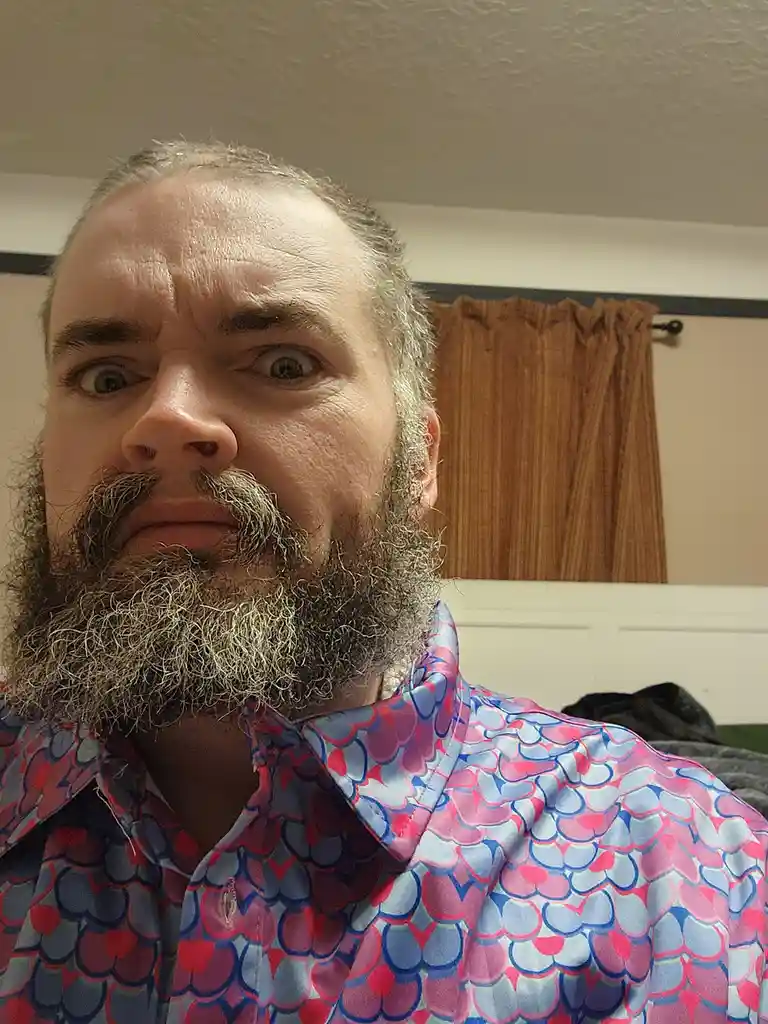 A man with a full beard and mustache stares directly at the camera with wide, startled eyes. He wears a brightly colored…