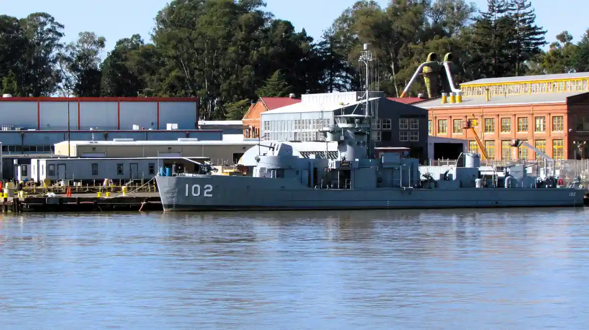 A gray naval vessel with the number 102 painted on its bow and stern is docked in calm water, with industrial buildings and…