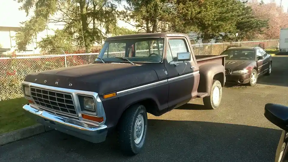 A dusty, dark purple 1970s Ford pickup truck is parked in a lot, with a maroon sedan visible behind it. The truck has a…