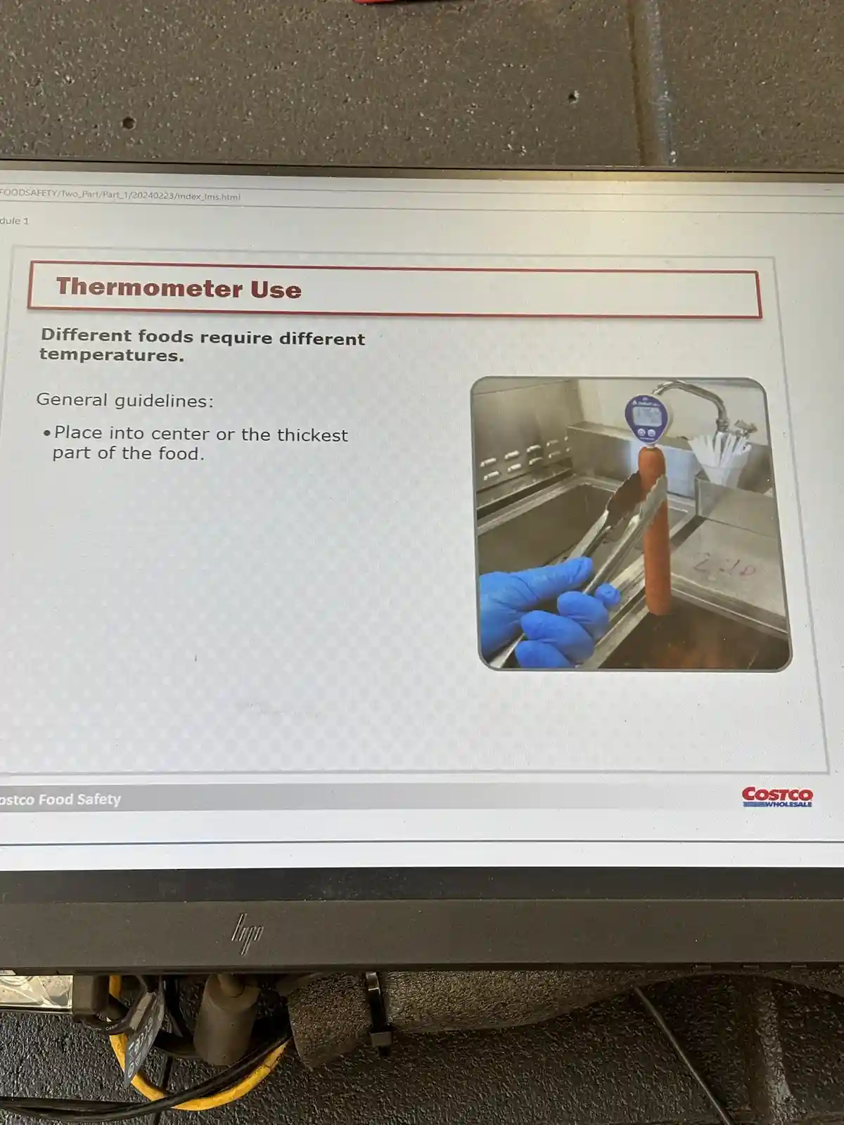 A computer monitor displays a Costco Food Safety training slide titled 'Thermometer Use,' which instructs users to place the…