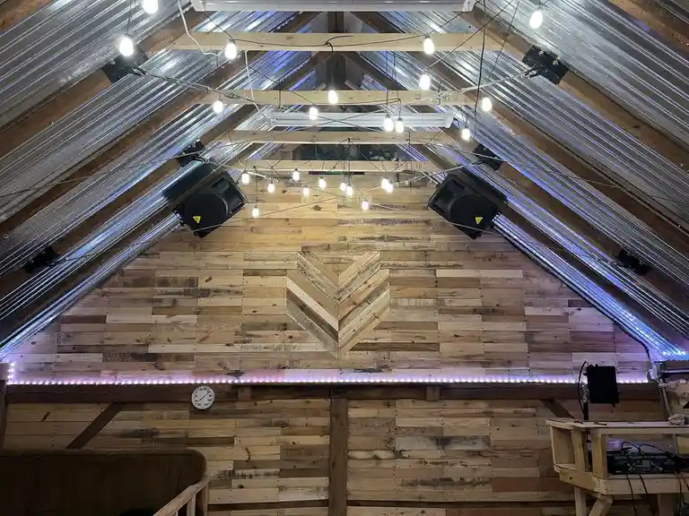 An attic room with a wooden wall and corrugated metal roof, featuring string lights, two large black speakers mounted on the…