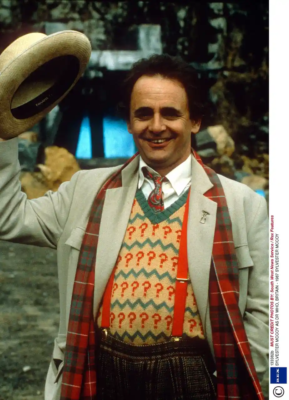 A man in a light grey suit, red plaid scarf, and a sweater covered in question marks raises a straw hat in a cheerful…
